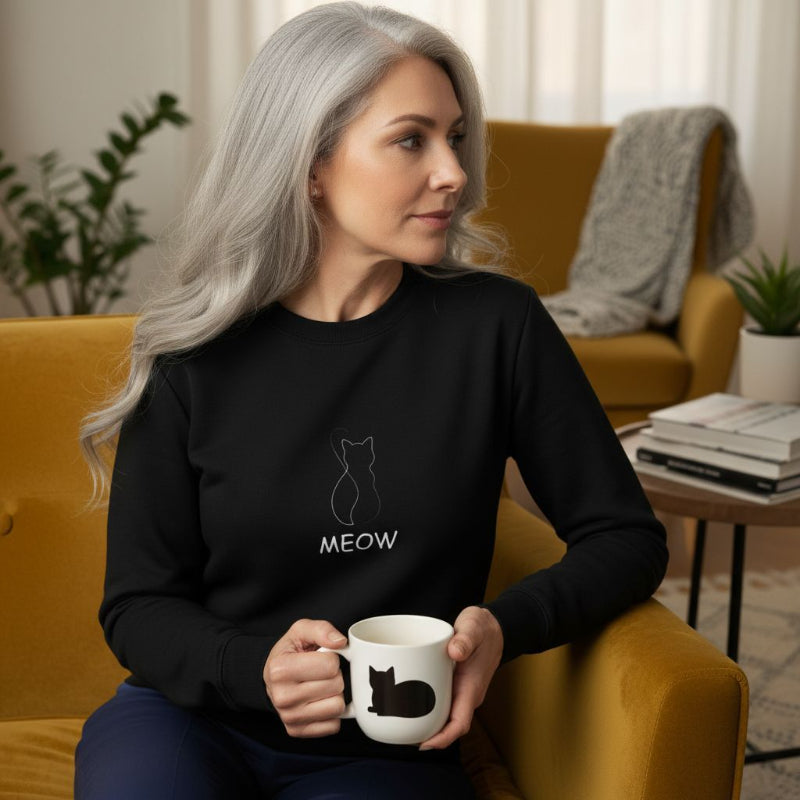 Woman sitting on a yellow couch holding a mug with a cat design, wearing a black sweatshirt with a cat icon and 'MEOW' text.
